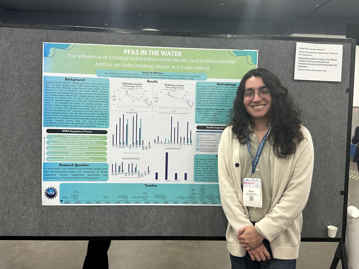 A college Fulbright student with log wavy hair and glasses wearing a beige cardigan stands in front of a research poster