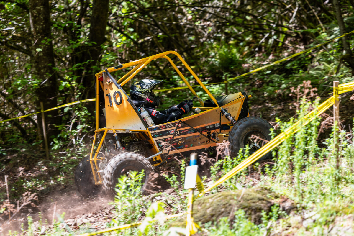 Off-road Baja car drives up hill with vegetation on either side.
