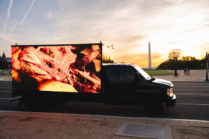 A truck with a large digital screen on its side displaying an abstract image travels past the Washington Monument at sunset.