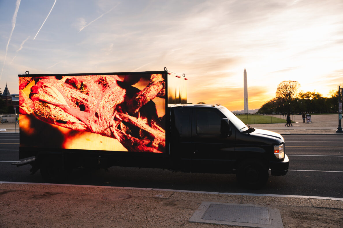 A truck with a large digital screen on its side displaying an abstract image travels past the Washington Monument at sunset.