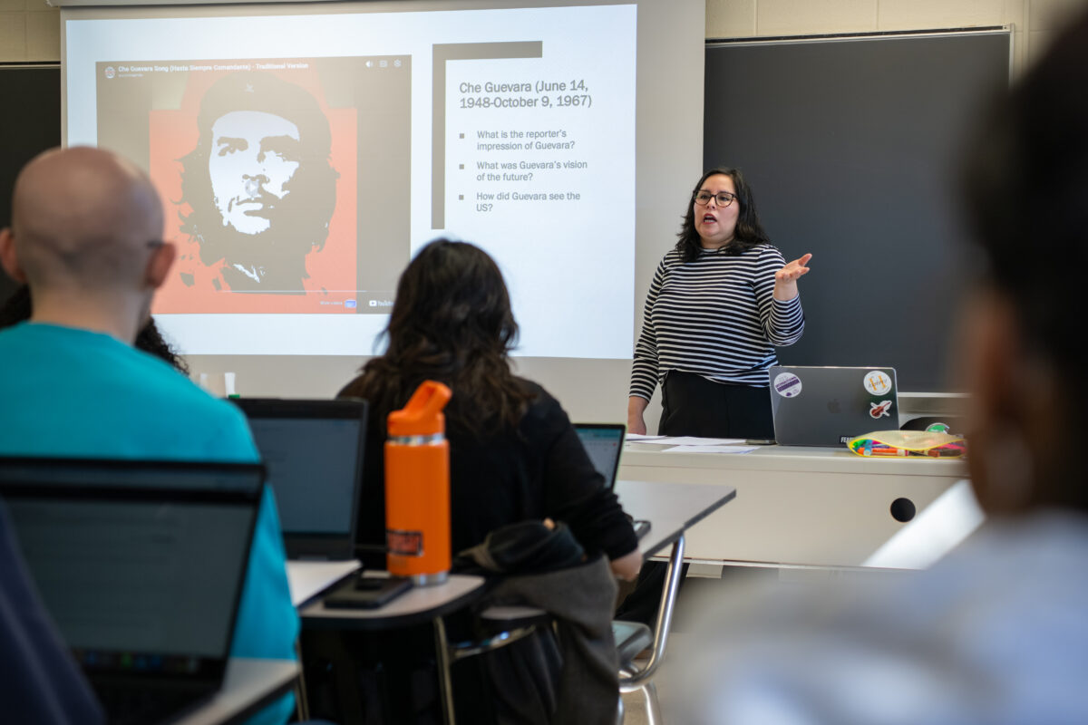 A college professor wearing a black and white stripped shirt and black pants stands in front of a classroom of students next to a projected black and white  image of Che Guevara on a red background wearing a black beret with a star 
