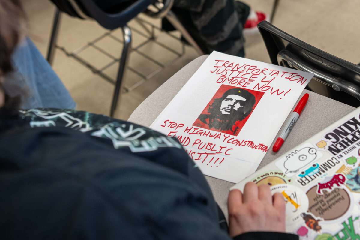A college student looks at a paper with a black and white headshot of a man wearing a black beret with a white star in a red background surrounded by words about transportation access