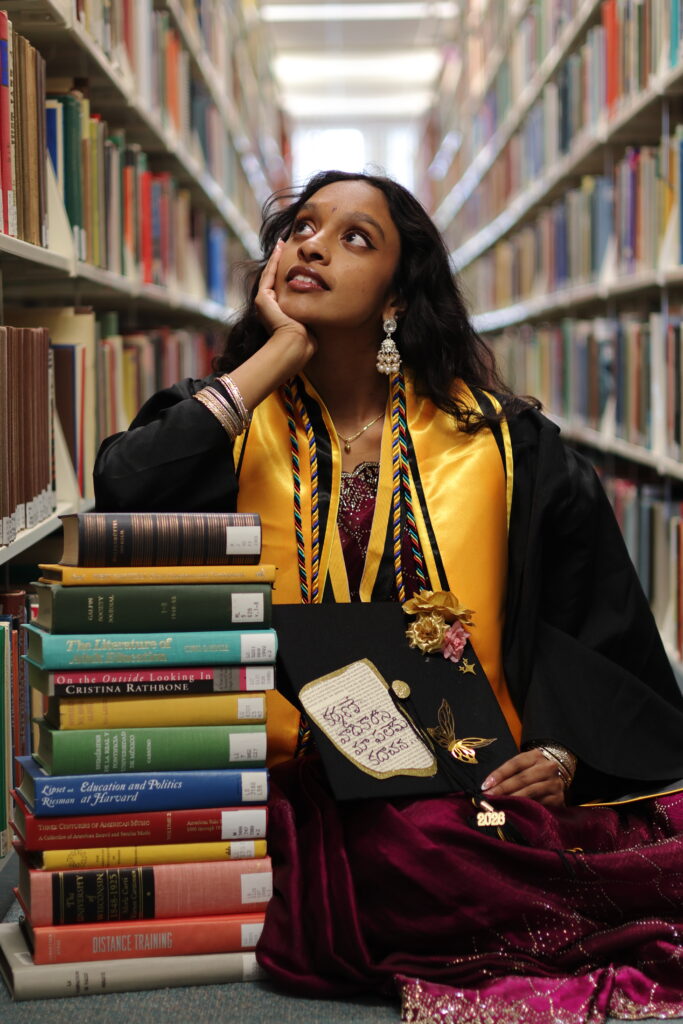 A college student wearing graduation regalia sits between two library shelfs leaning on a stack of books