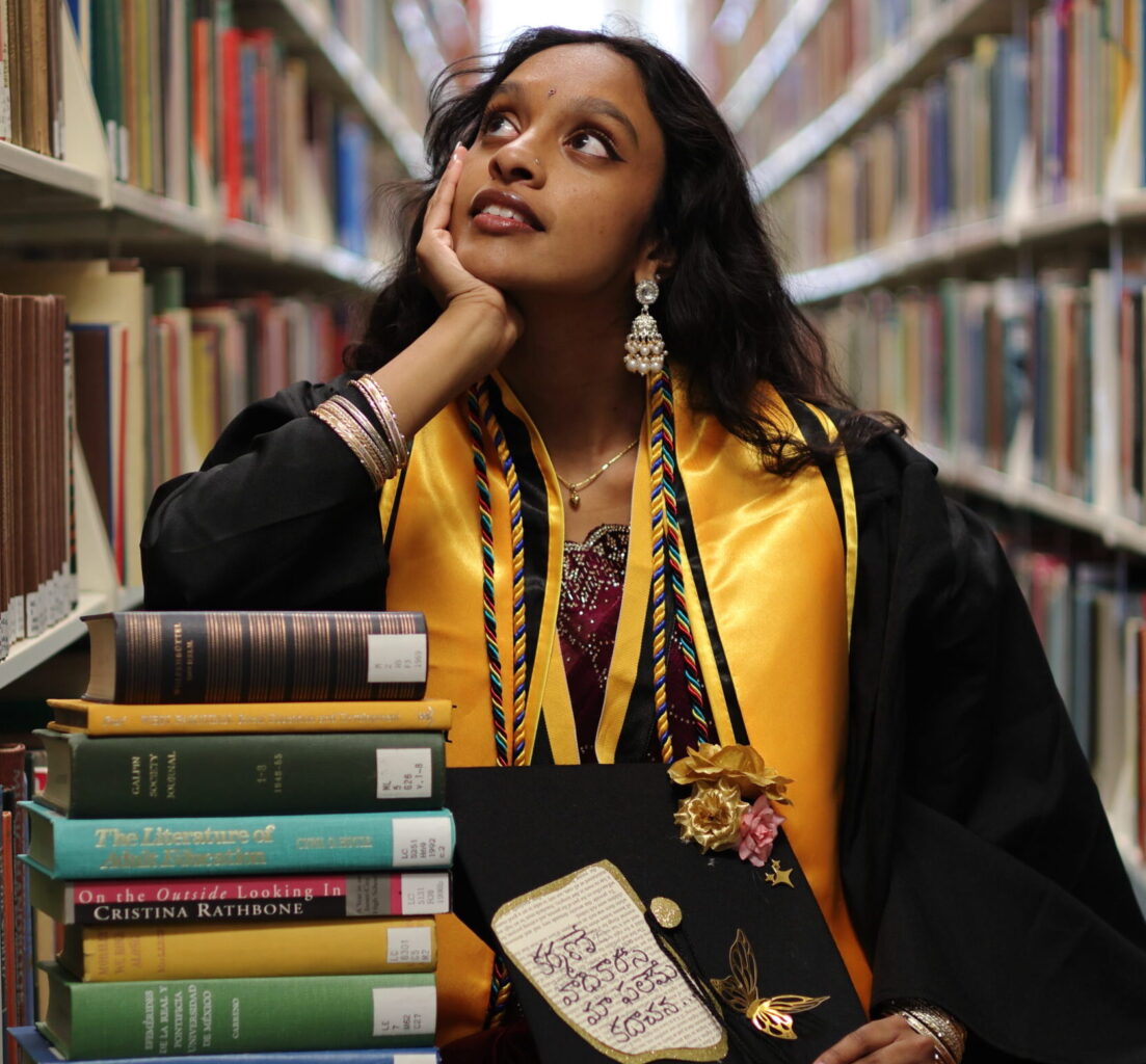 A college student wearing graduation regalia sits between two library shelfs leaning on a stack of books