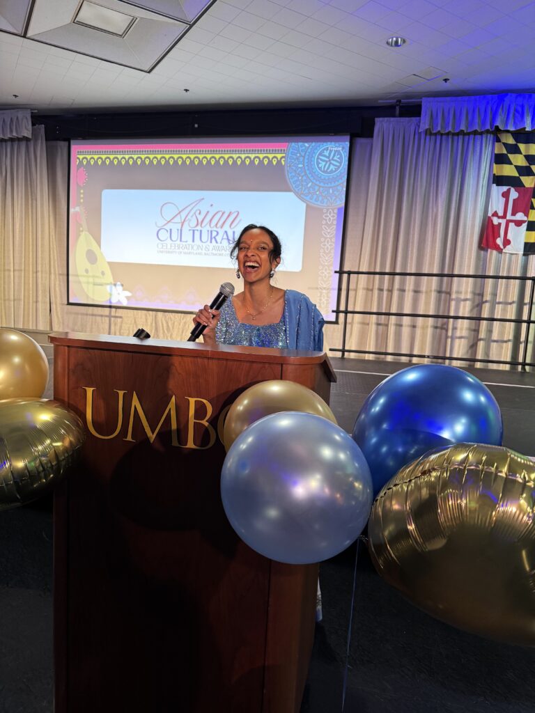 A Fulbright student wearing a purple dress stands behind a podium holding a mic