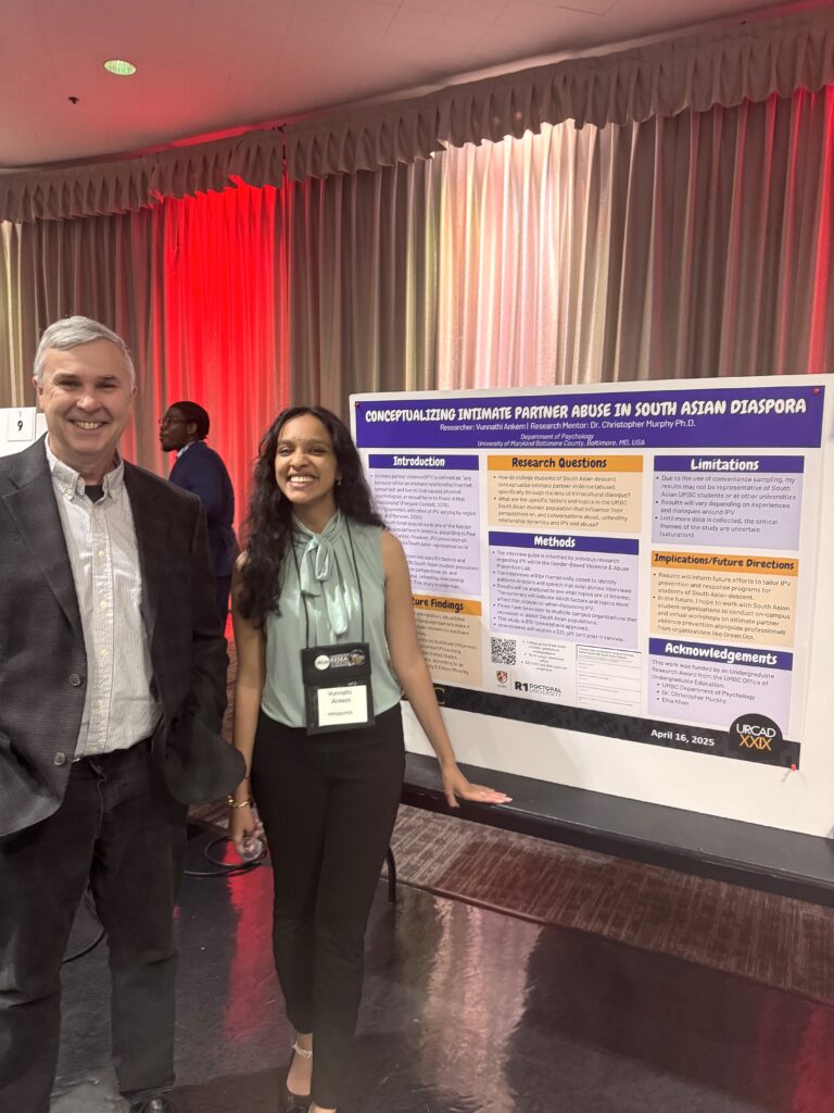 A college professor wearing a grey suit stands next a student Fulbright wearing black pants and a mint blouse stand in front of a research poster