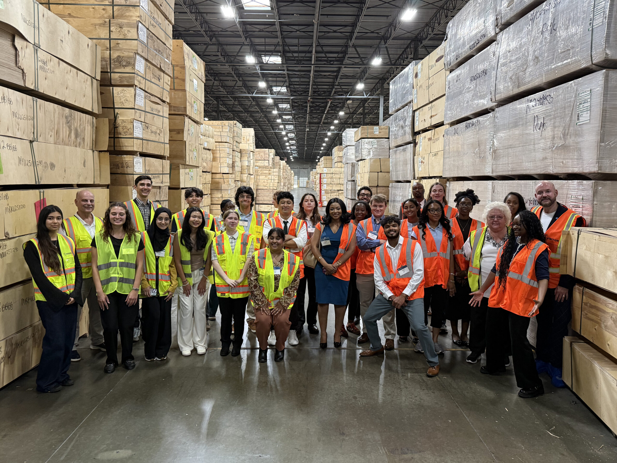 a group of people in safety vests stands in a warehouse full of crates