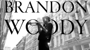 A person plays a trumpet in a city street. The text reads "BRANDON WOODY."