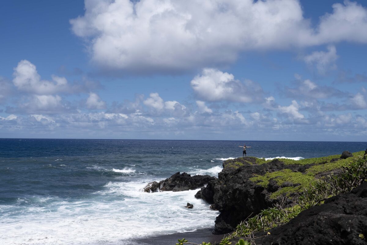 Noah Reichardt stands on a rocky point, arms outstretched, with crashing weaves, blue sky, and puffy clouds surrounding him