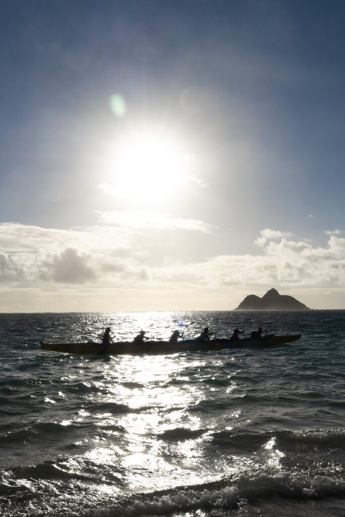 six people in a boat rowing, an island in the background, as the sun rises