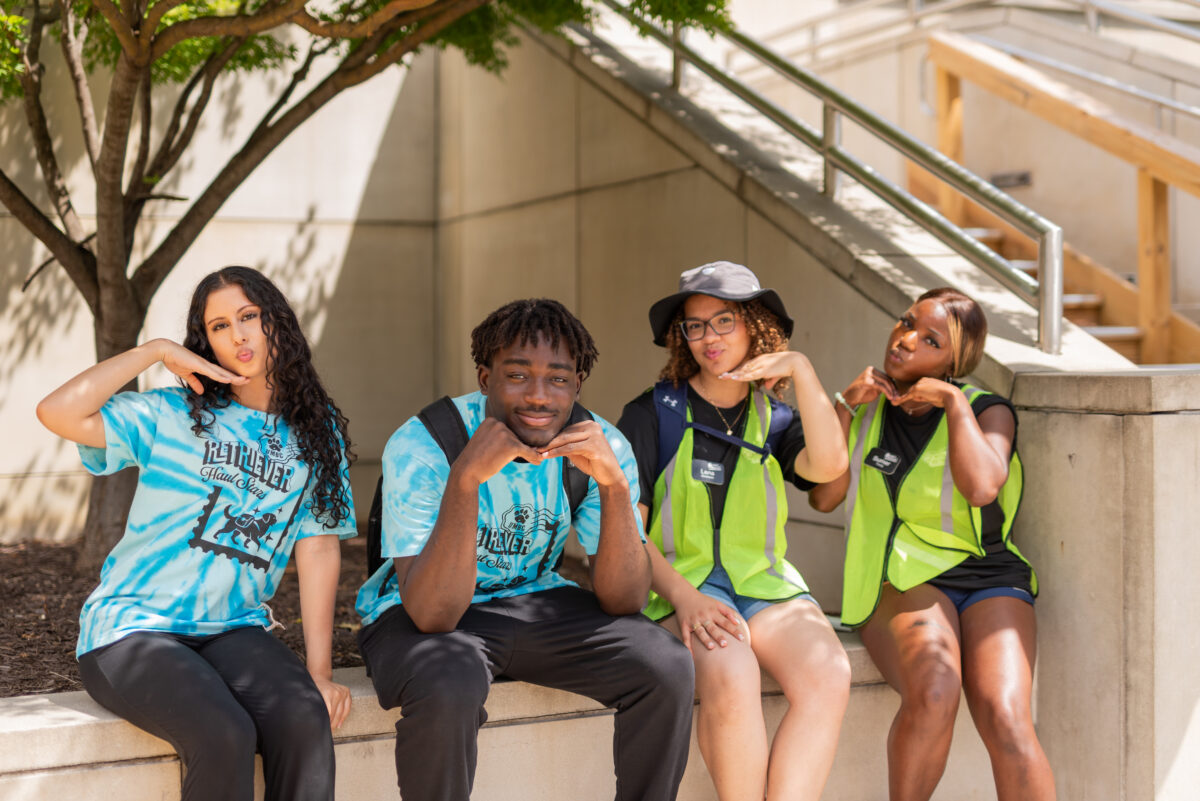 four students sit in the shade, they wear brightly colored shirts and safety vests to show that they're here to help new students move