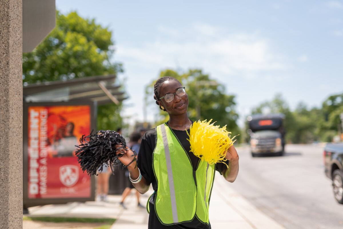 someone in a yellow safety vests waves a pom pom to welcome new students to campus