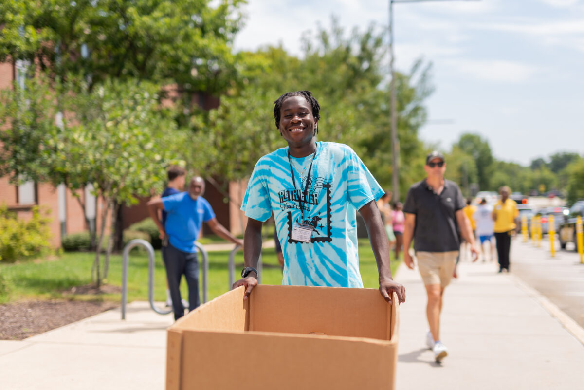 a student in a bright blue tie-dyed shirt pushes a moving container