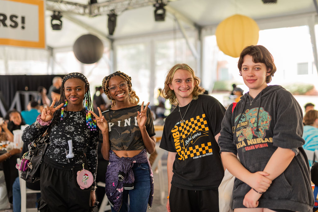 four students gather under a large outdoor tent and pose together, giving the peace sign and large smiles