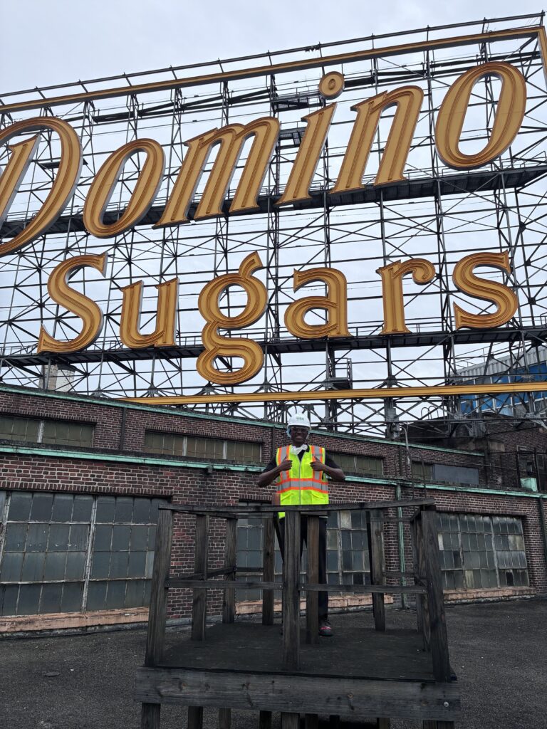 Pelumni Enilolobo, wearing a reflective safety vest, outside under a large Domino Sugars sign.