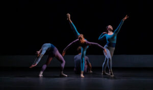 Four dancers perform a contemporary dance routine on a dark stage.