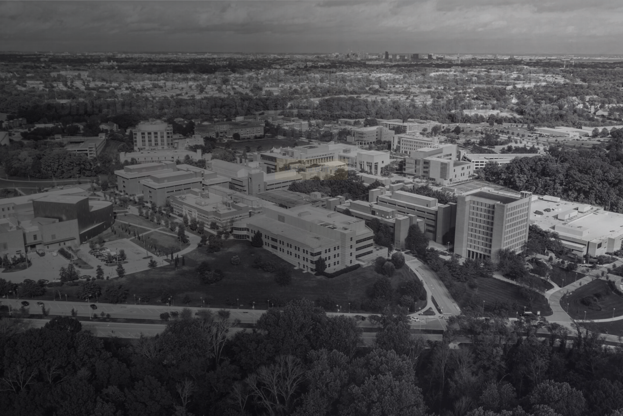 Aerial view of the UMBC campus with multiple academic buildings, parking lots, and surrounding trees under a cloudy sky in black and white.