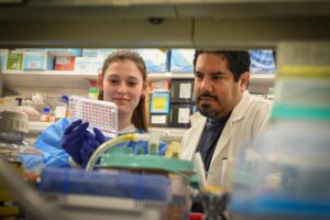 Magdalen Ruth and her mentor work at a lab bench in gloves and lab coats; she's holding a well plate