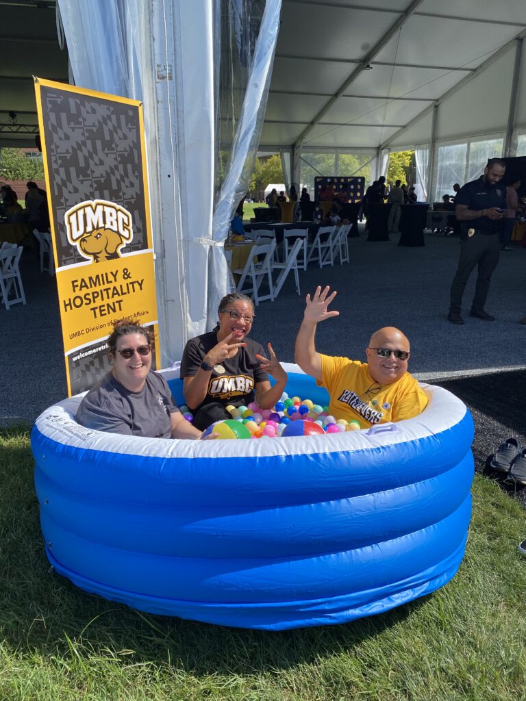 three adults are in a child-size ball pit with a sign behind them that says UMBC Family and Hospitality Tent