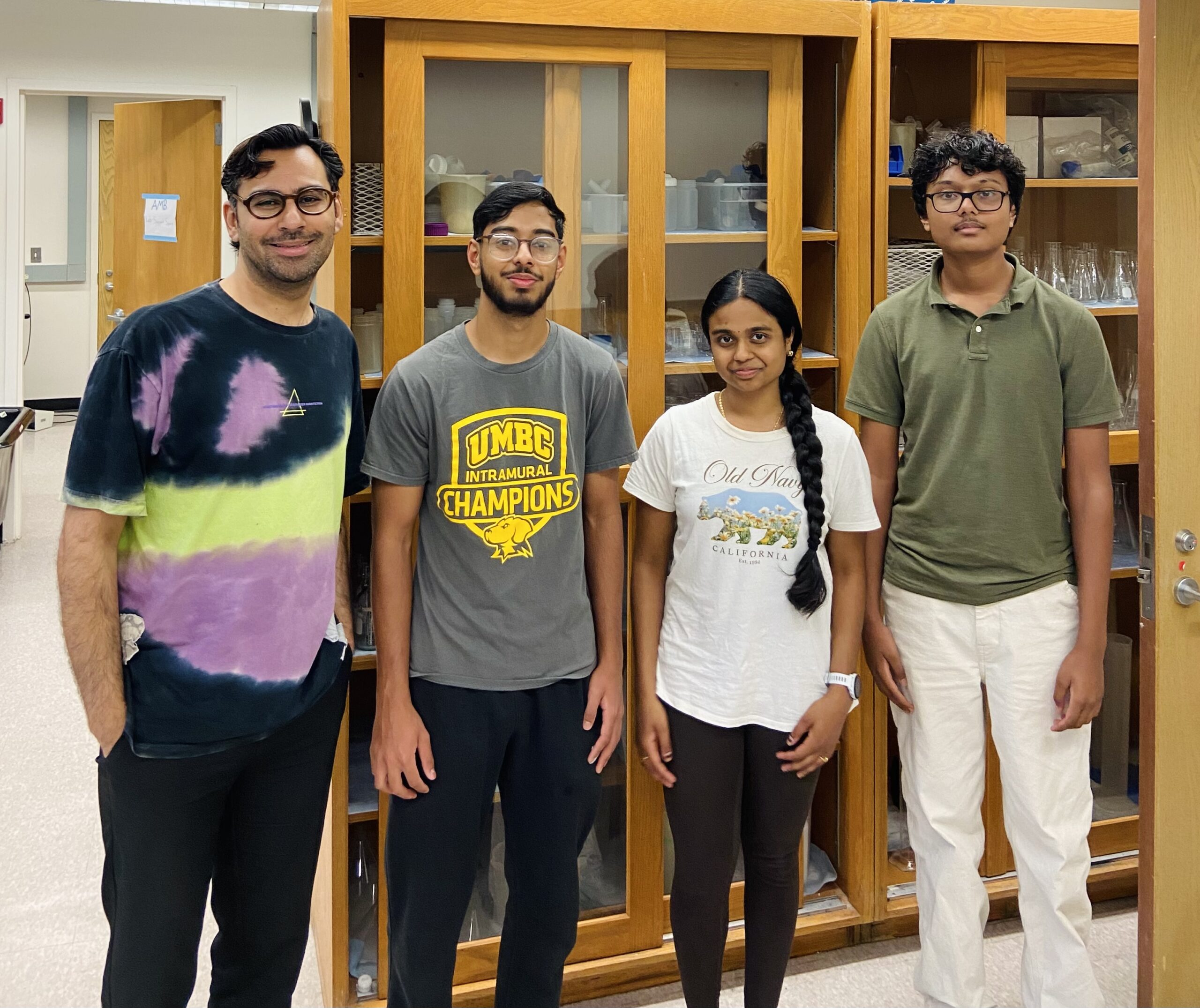 group photo of Mayank Chugh with his three summer student researchers in front of glass cabinets in a lab