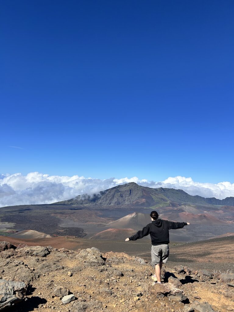 Morgan Robbins hikes, arms outstretched, along a rocky landscape with a large mountain in the background