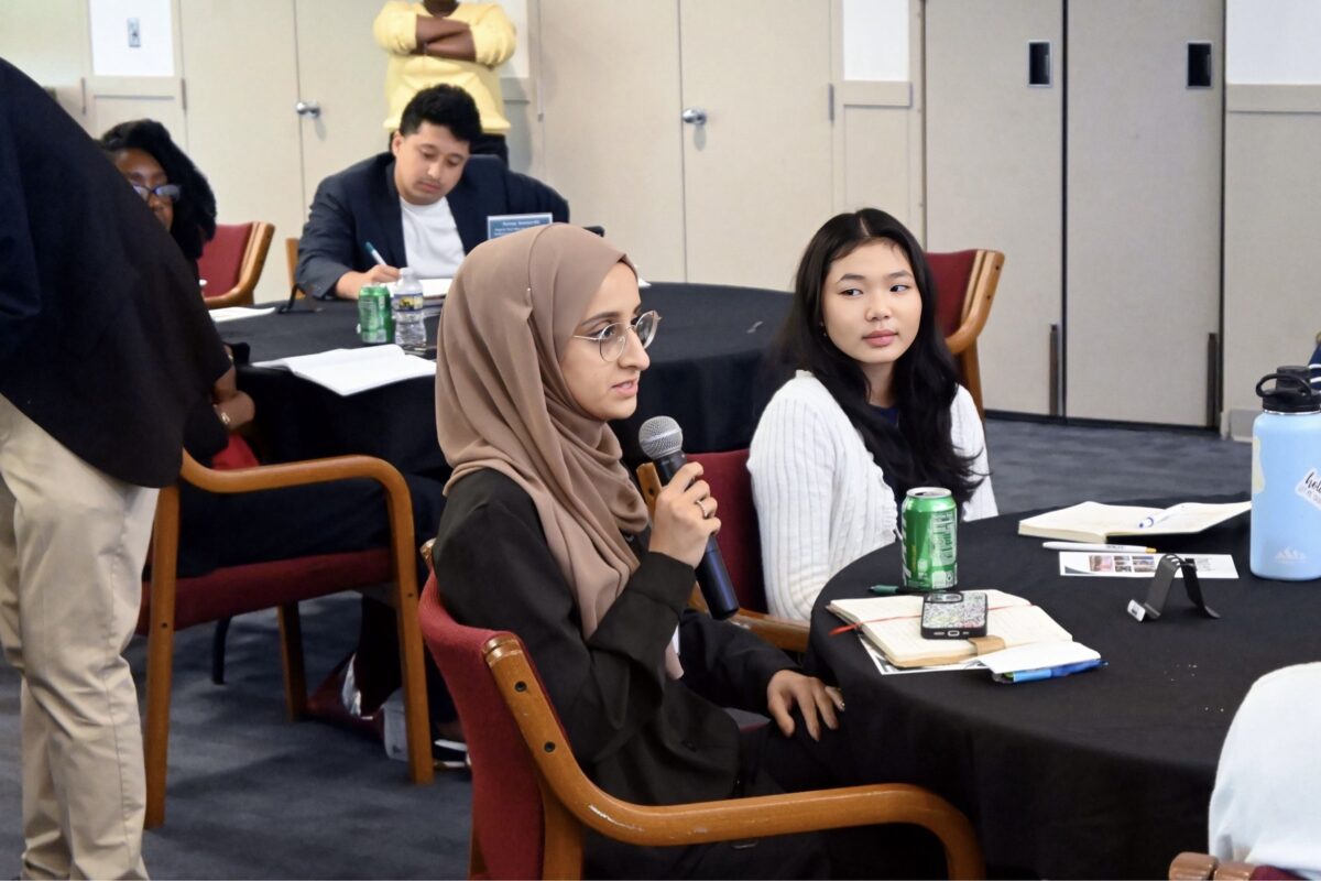 a woman in a tan headscarf sits at a table with others while talking on a microphone