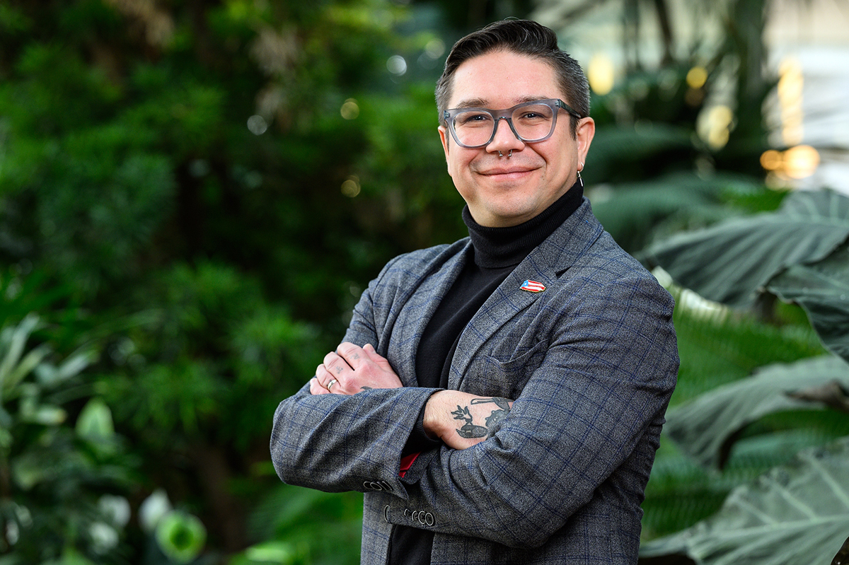 Headshot of a person wearing glasses and a blazer with arms crossed, standing in front of lush green plants.