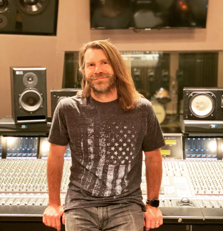 A person sits at a large audio mixing console in a recording studio, with monitors and equipment visible in the background.