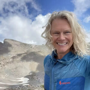 A person smiles while standing on a mountain slope with rocky terrain and patches of snow.