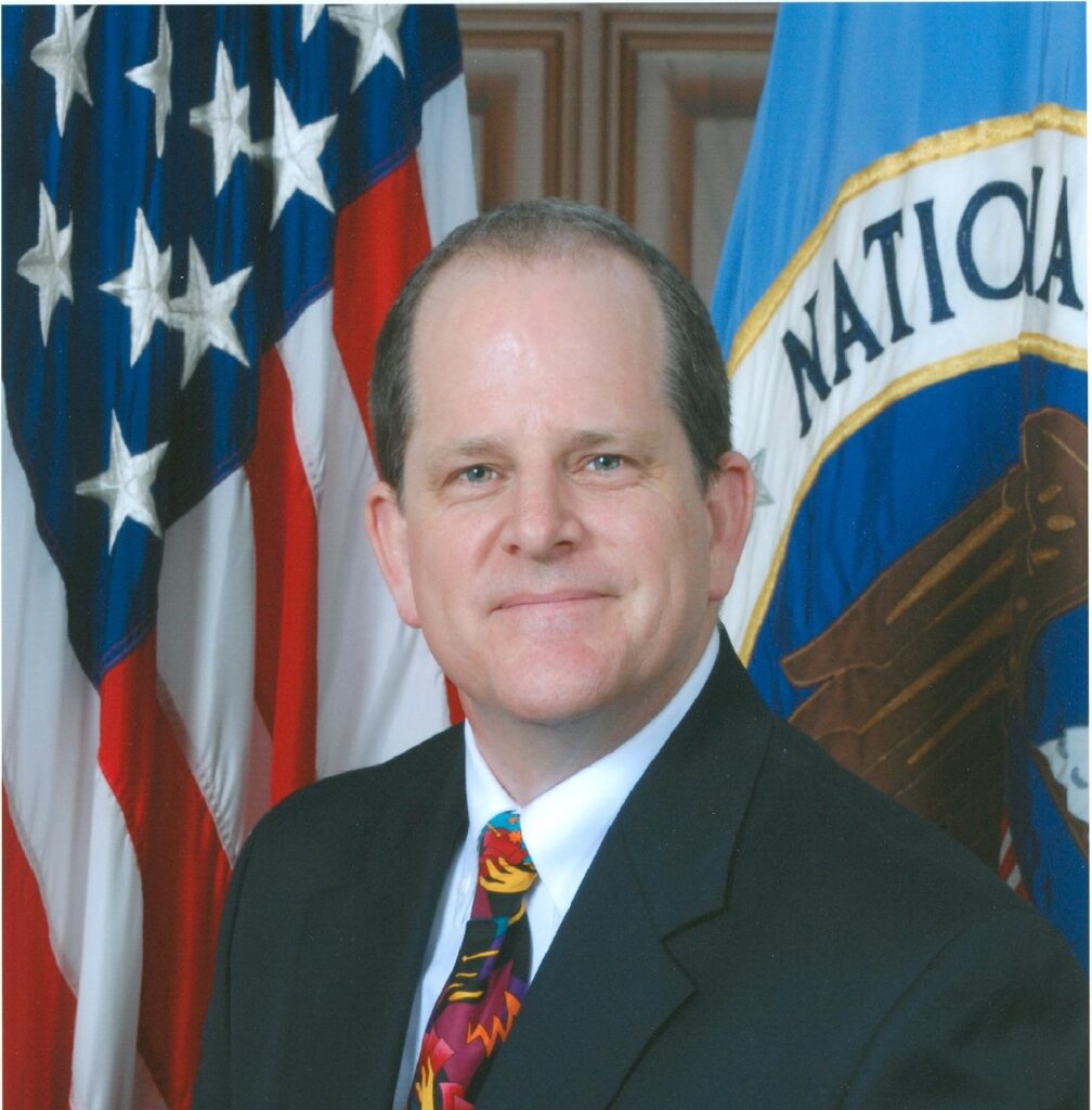 Head shot of David Mountains in a suit and colorful tie in front of a U.S. flag a NSA flag.