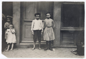 Four children standing in front of a doorway. A young boy stands barefoot, and three other children wearing simple clothes stand nearby.