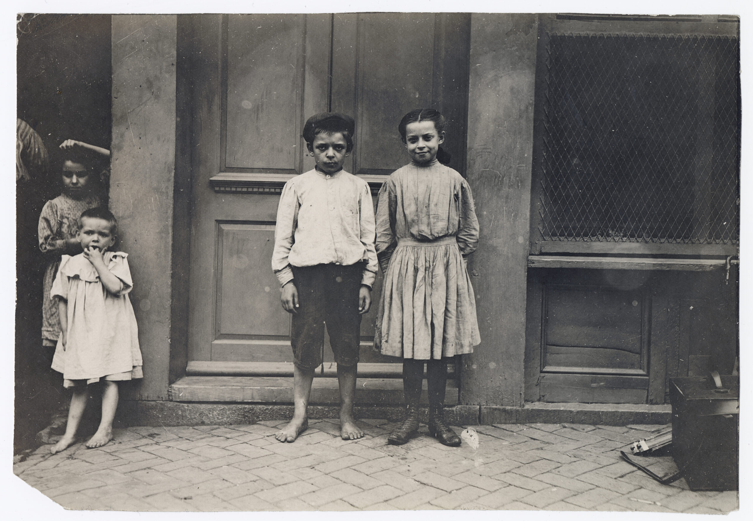 Four children standing in front of a doorway. A young boy stands barefoot, and three other children wearing simple clothes stand nearby.