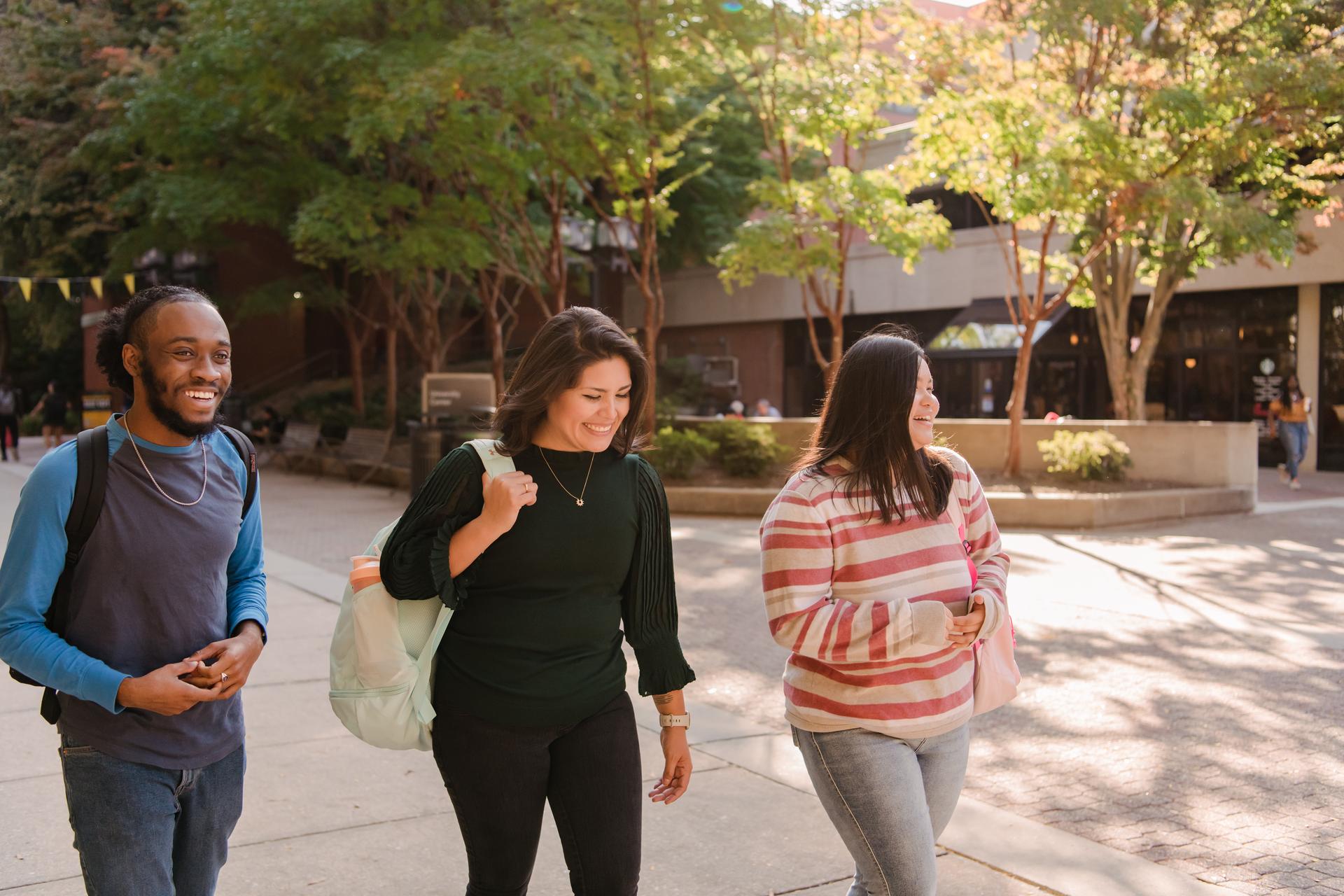 Three students with backpacks walk outdoors on campus while talking and smiling.