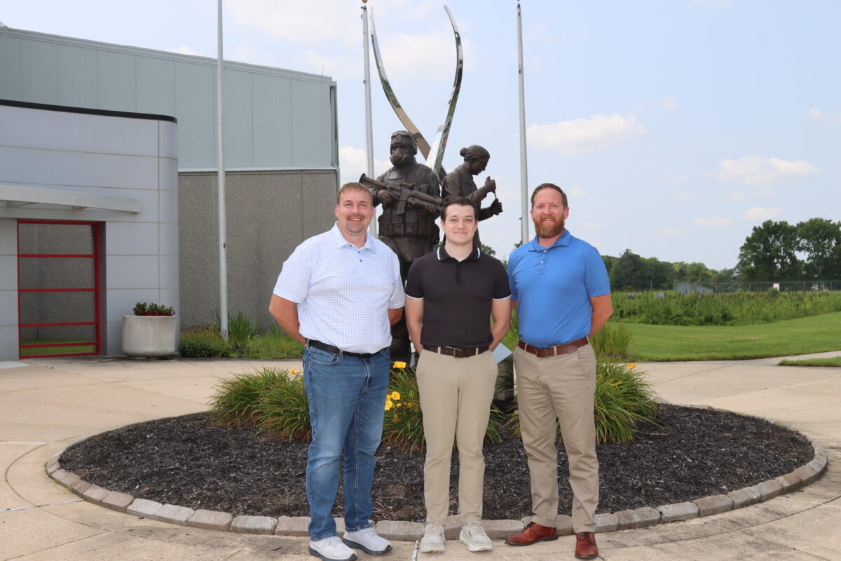 Jon Sampson, Mark Harrison, and Ed Parshley stand in front of a bronze statue of two figures holding equipment.