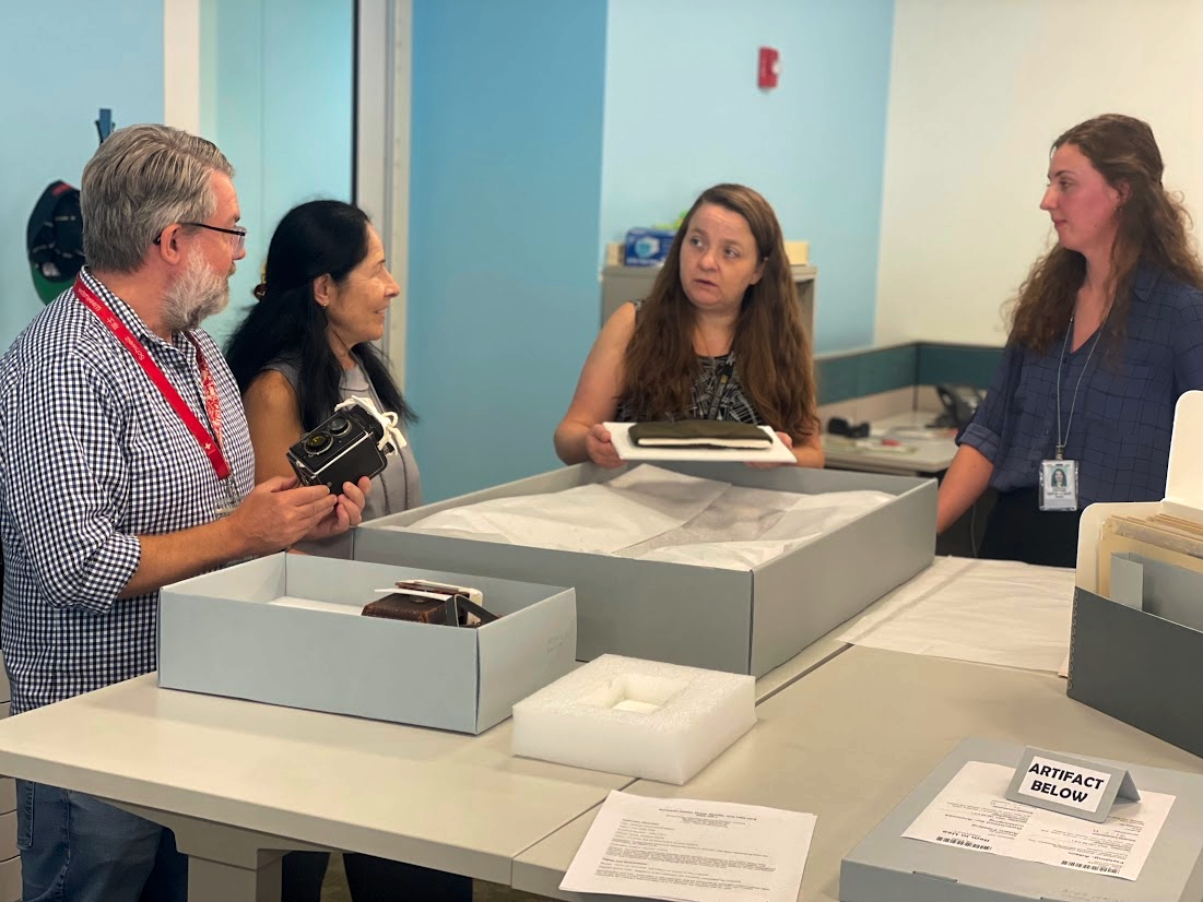 Four museum archivist stand around a table with open archival boxes that hold a WWII soldier hat and a camera.