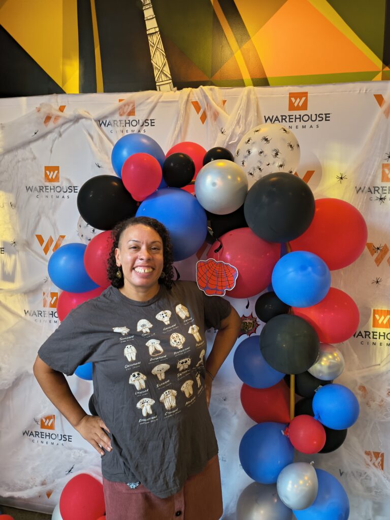 Mercedes Burns standing in front of a display of red, black, and blue balloons, wearing a t-shirt with spiders on it
