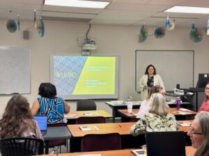 Diane Zeenny Ghorayeb, a presenter stands in front of a college classroom next to a projection screen displaying a black and gold presentation slide to lead an international immigration training on visas