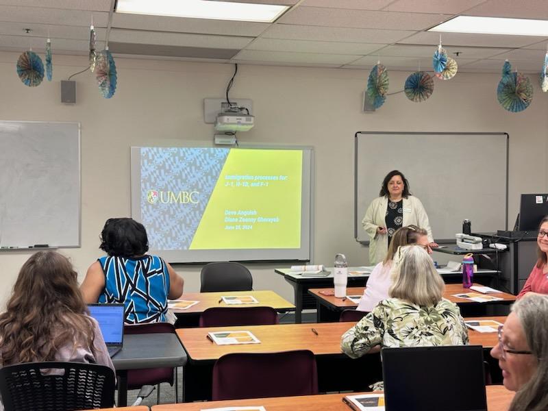 Diane Zeenny Ghorayeb, a presenter stands in front of a college classroom next to a projection screen displaying a black and gold presentation slide to lead an international immigration training on visas