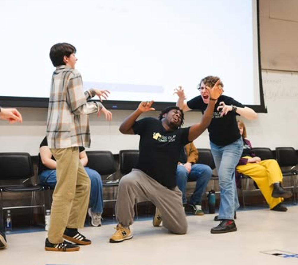 Three college students practice improve comedy with one student kneeling down with their arms in the air and the other two hover above him with their hands shaped like claws