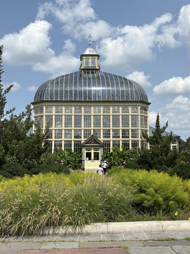 The glass dome of a conservatory in Baltimore City surrounded by gardens and trees, with a few people walking toward the entrance.