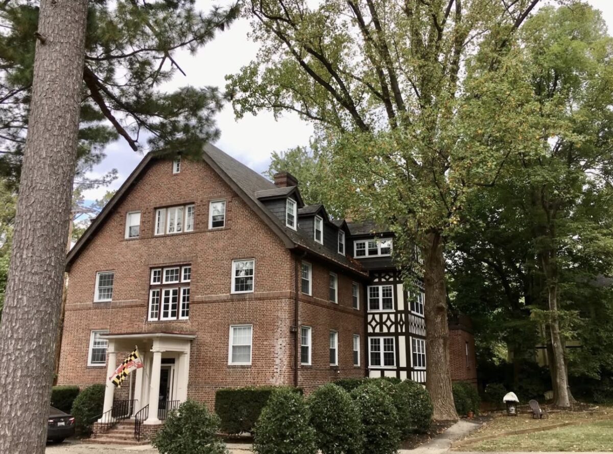 A brick building with a Maryland state flag displayed at the entrance, surrounded by large green trees.