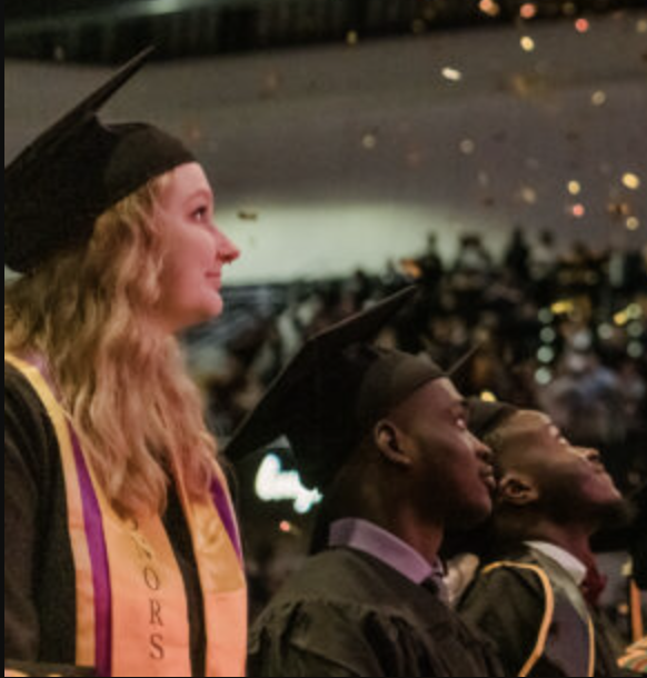 Students in academic regalia look up at falling confetti during a commencement ceremony.