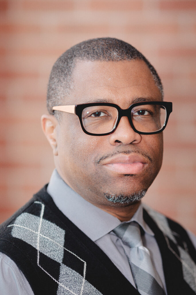 headshot of a man, working on a program for incarcerated students in Maryland, with short grey hair, black rimmed glasses, a grey dress shirt and tie, and a black, grey, and white argyle sweater, Shriver Center