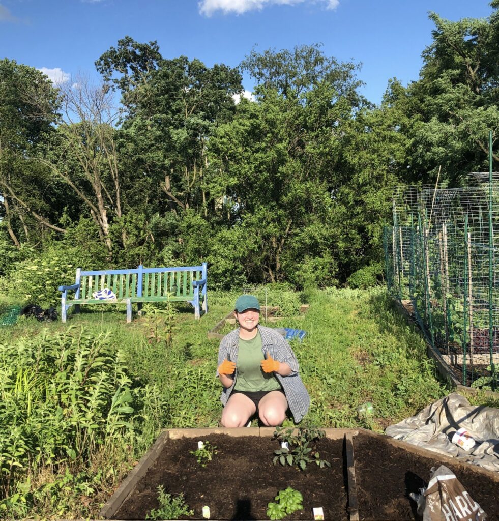A college student wearing a baseball hat and gardening gloves poses for a picture kneeling in front of her garden plot