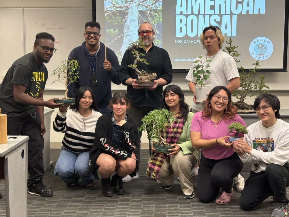 Nine people stand together in a classroom holding bonsai trees, in the background there is a projection screen with a slide with the words American Bonsai