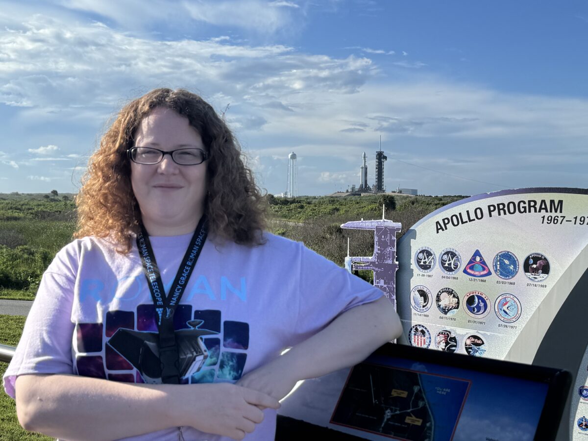 Rebekah Hounsell stands next to an informational placard that reads "Apollo Program," with the SpaceX Falcon Heavy rocket that would carry the Roman telescope into space visible in the background at a distance.