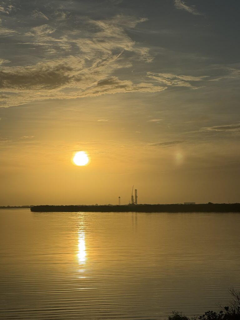 sunset approaches as the  sun hangs low in a golden sky, with the rocket visible at a great distance across water 