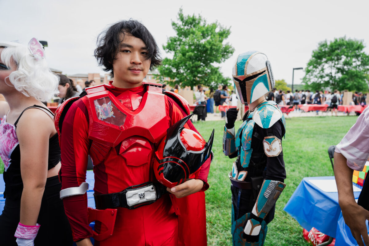 Two college students face the camera. One is wearing a red super hero costume and the other is wearing a teal super hero costume