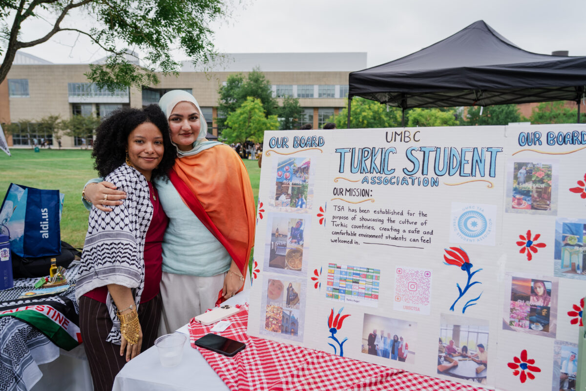 Two college students hug each other as they stand next to a presentation display board with information about Turkic Student Association