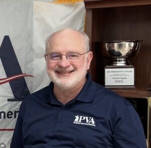 Headshot of Tim Besse wearing a polo shirt with the PVA logo, smiling in front of a shelf containing a trophy.